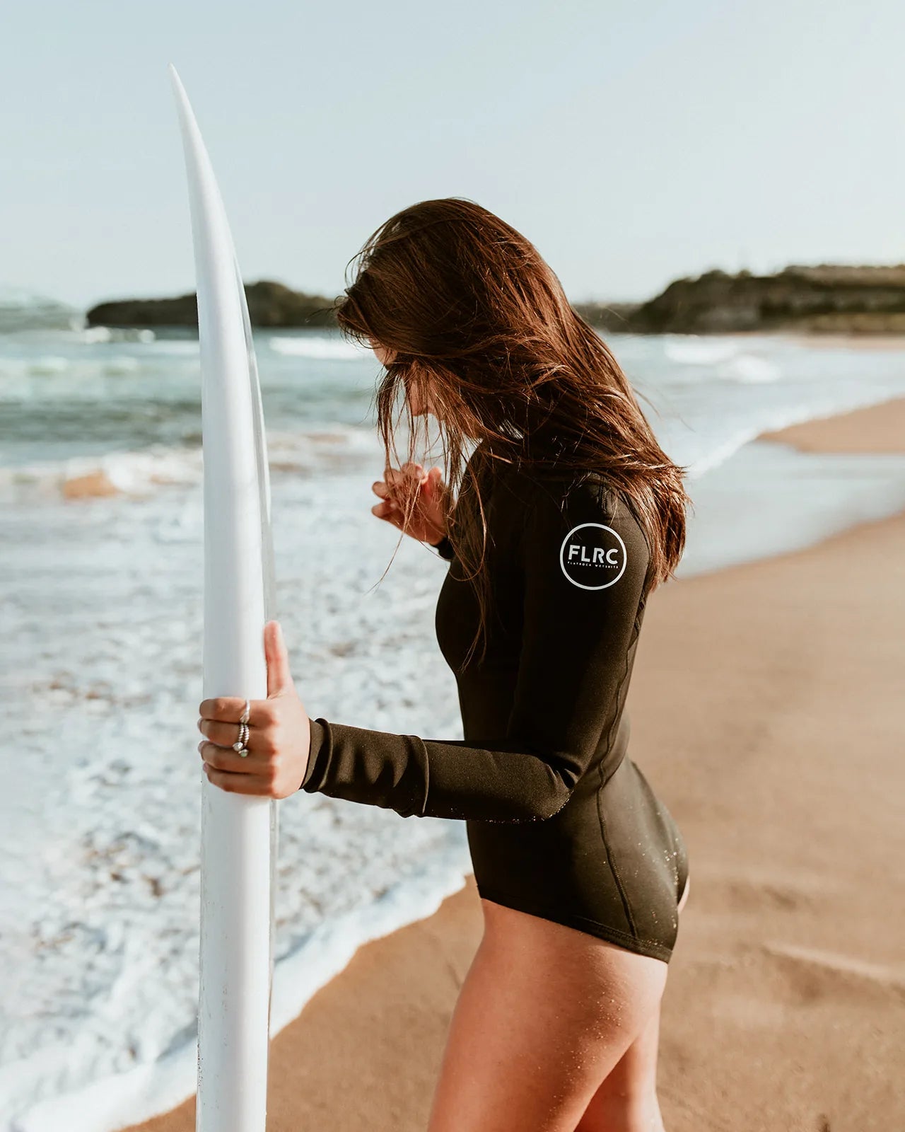 Woman wearing black FLRC 1.5mm long sleeve spring suit with white logo, side view holding surfboard on beach.