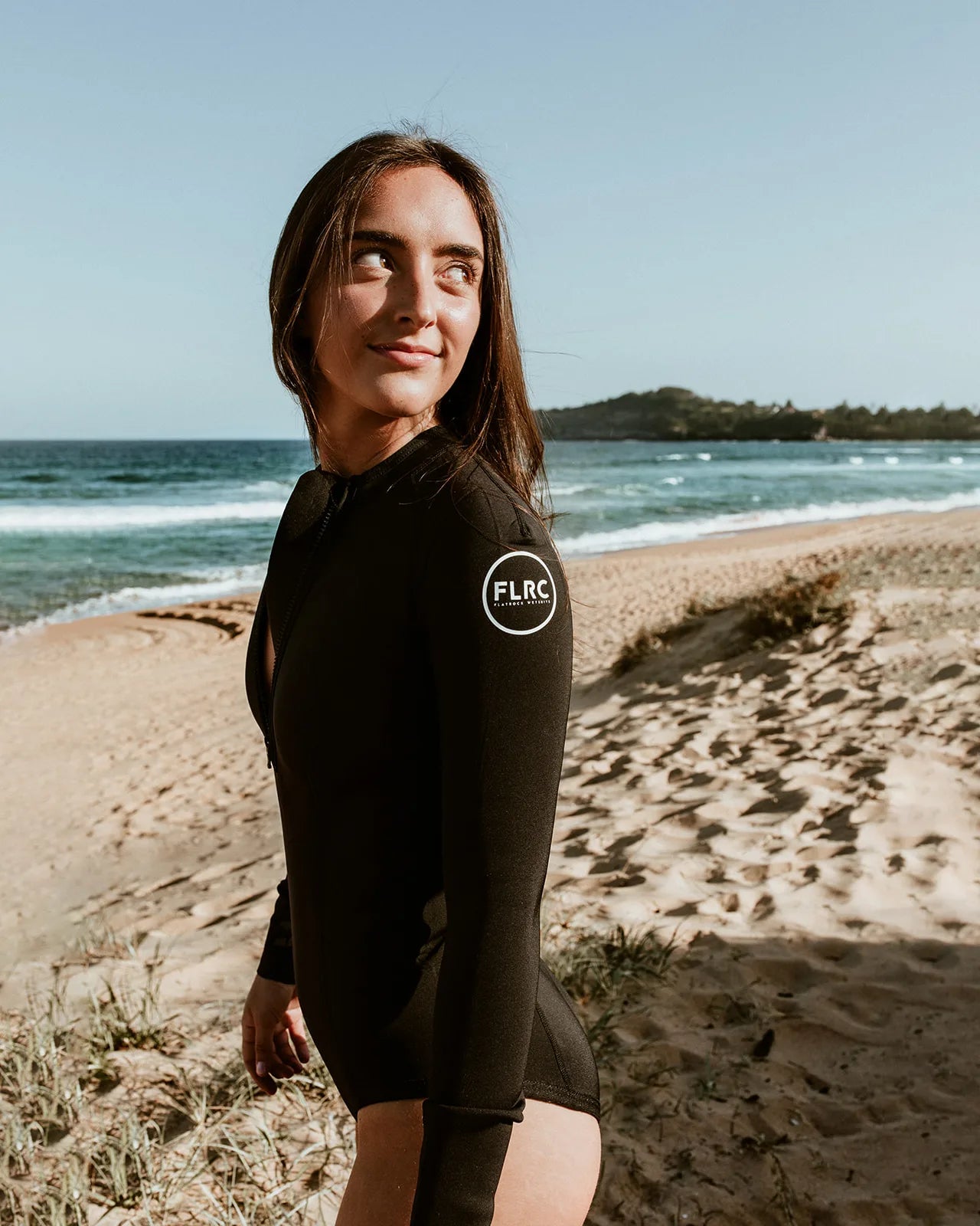 Woman wearing black FLRC 1.5mm long sleeve spring suit with white logo, side view on beach.