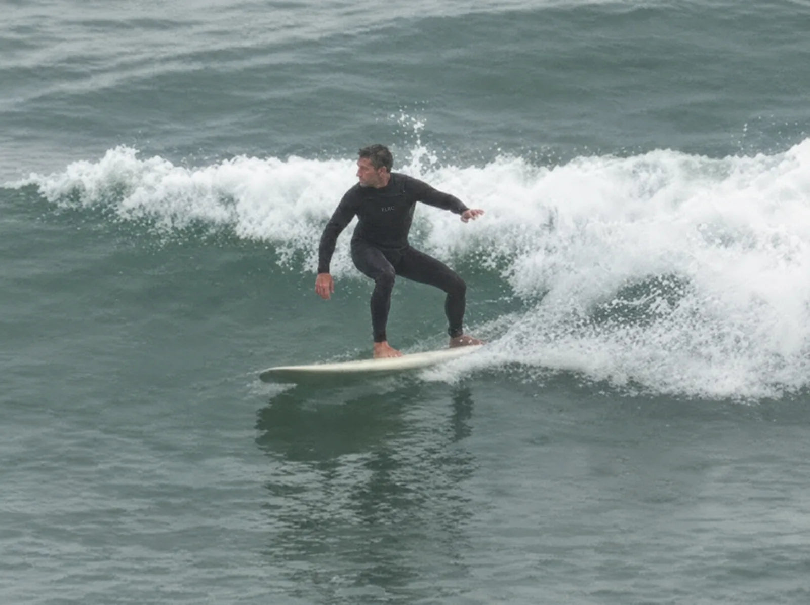 Surfer trimming along a small grey wave on a longboard in the V3 steamer, shot from the beach