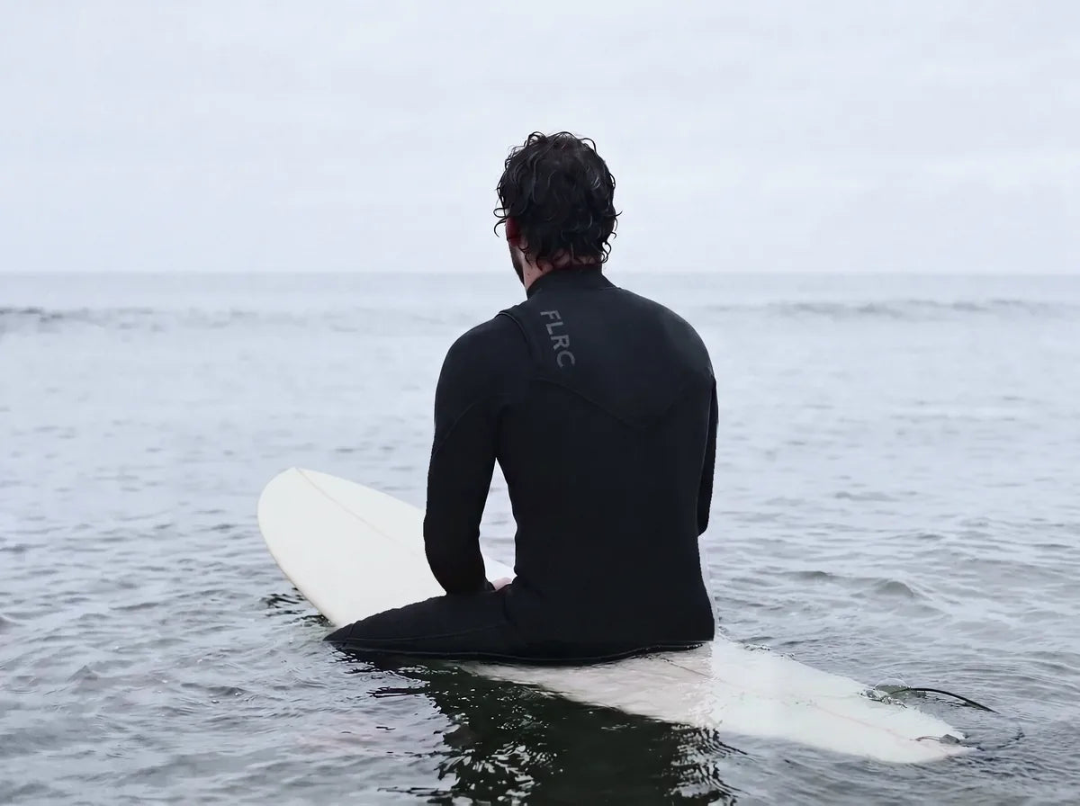 Surfer sitting astride a longboard on flat grey water, back to camera, FLRC across the shoulders