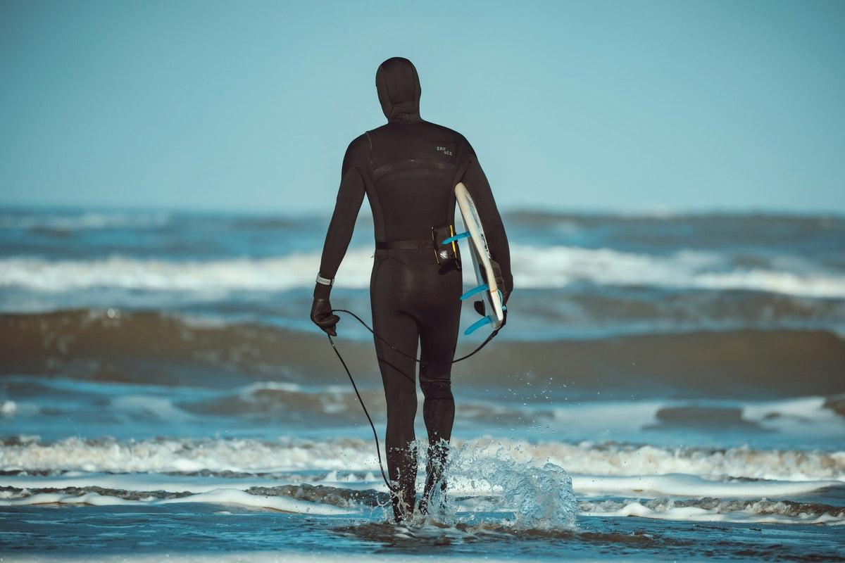 Surfer in a hooded full wetsuit wading into a cold sea with his board