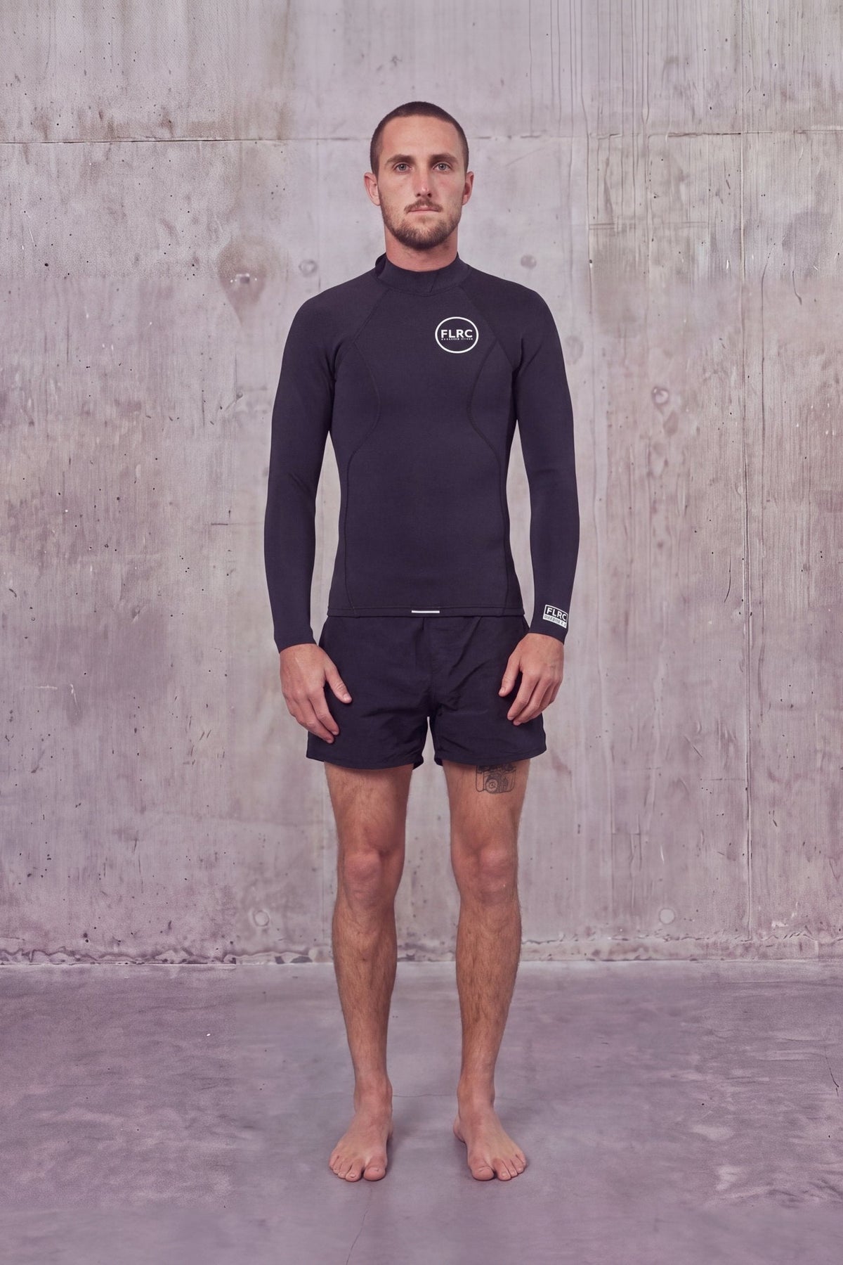 Man in Men's 1.5mm Wetsuit Top V1 with white FLRC chest logo, front view against a concrete wall
