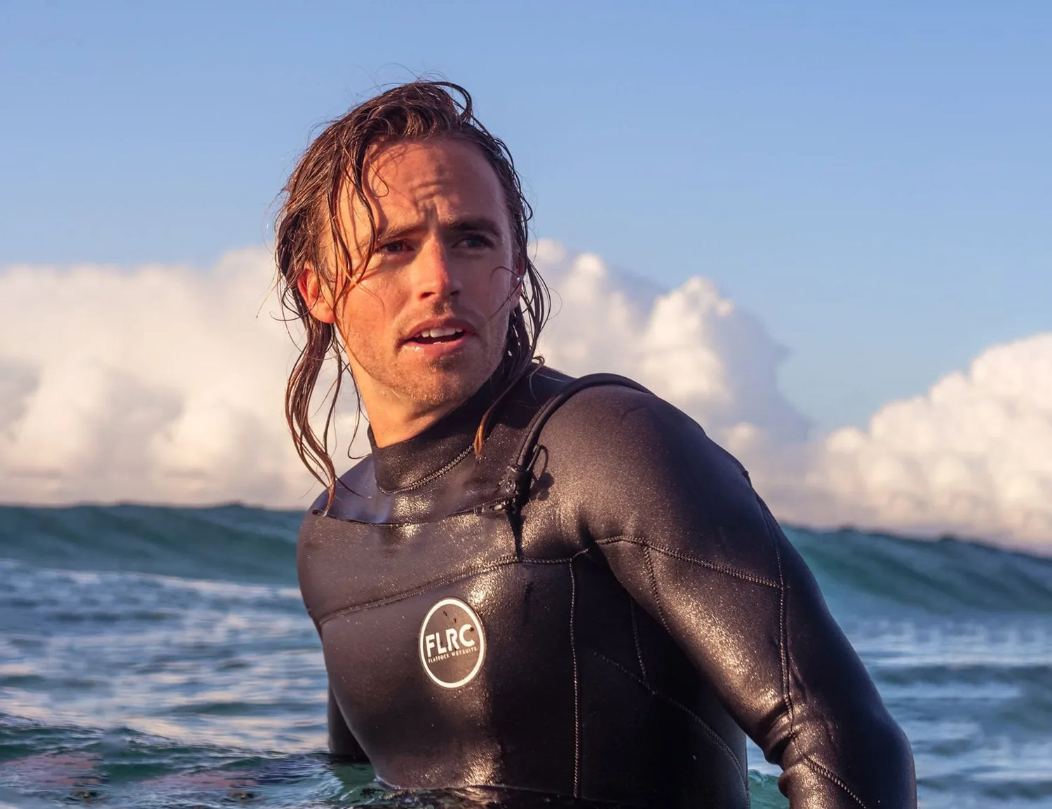 Man wearing black FLRC 3/2mm steamer wetsuit with white logo (2019 design), sitting in water, back view.