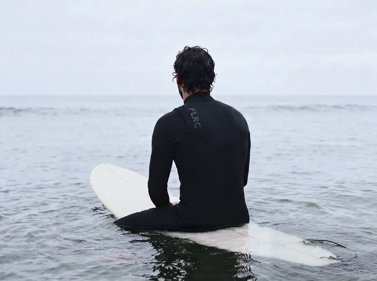 Surfer seen from behind, sitting on a surfboard in the water in a black FLRC wetsuit