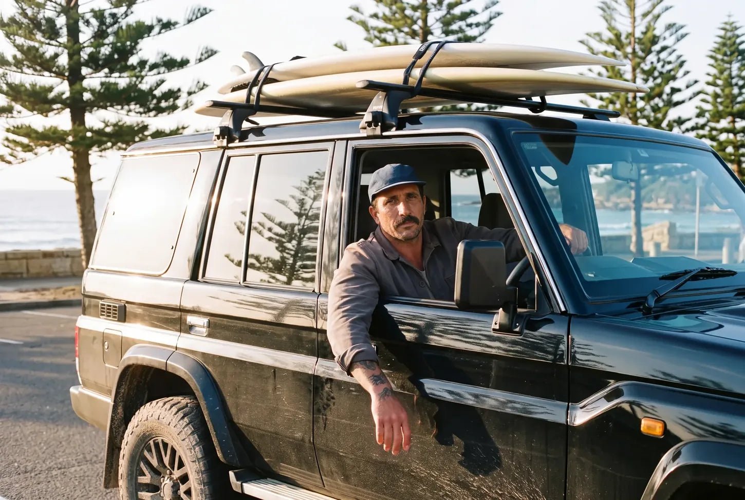 Surfer leaning out of a black LandCruiser with boards on the roof by the beach