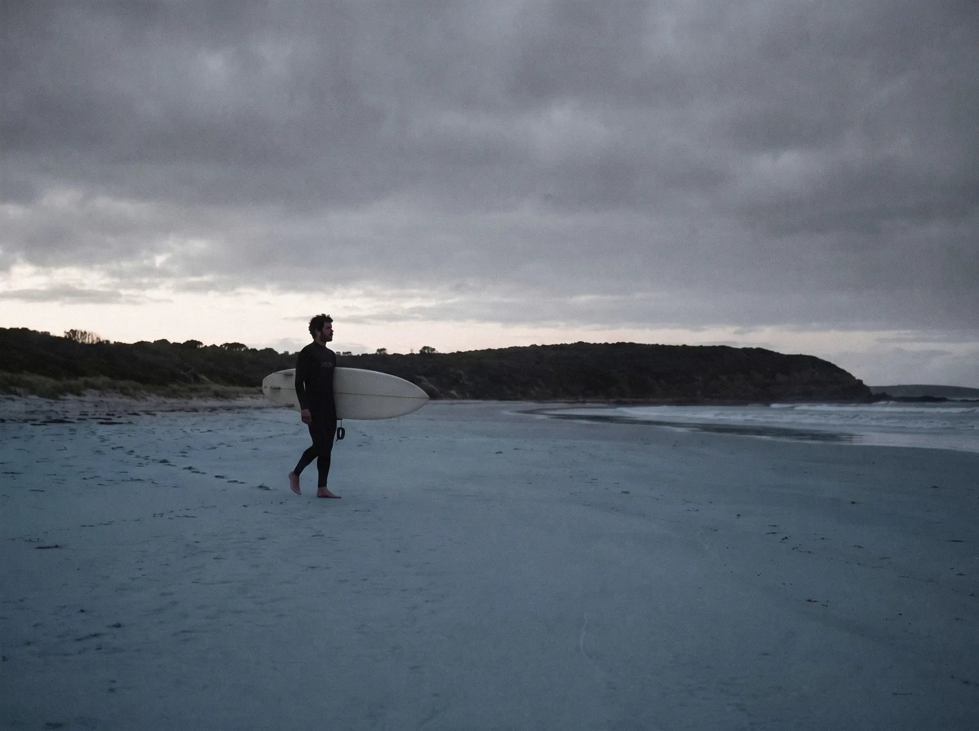 Surfer walking the beach at dusk with a board under his arm, headland behind