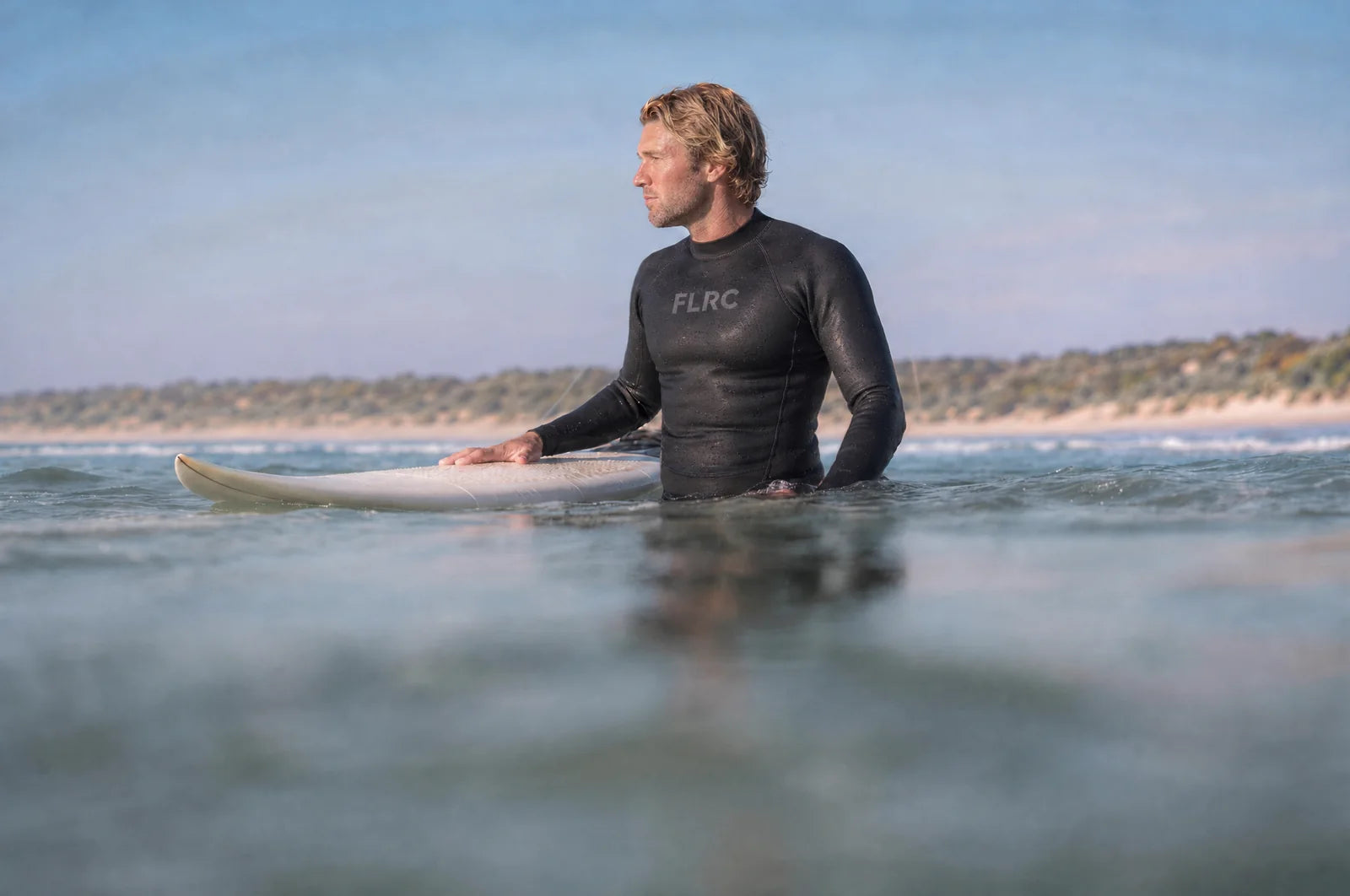 Surfer sitting on a longboard in the Bronte wetsuit top, a long Australian beach and low dunes behind him