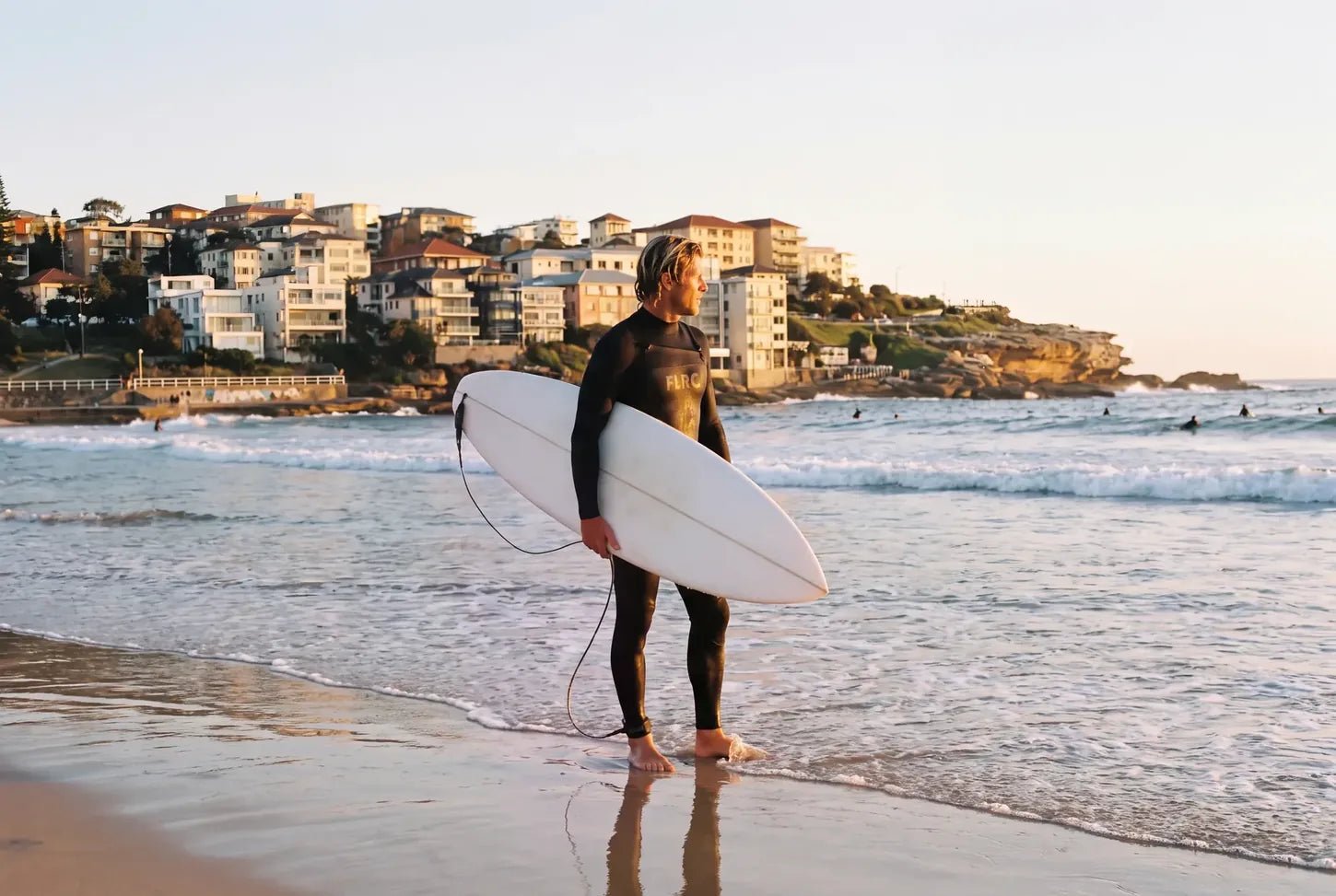 Surfer in a Flatrock steamer holding a board at the water's edge, Bondi at sunrise