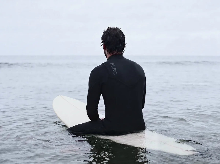 Surfer sitting astride a longboard on flat grey water, back to camera, FLRC across the shoulders