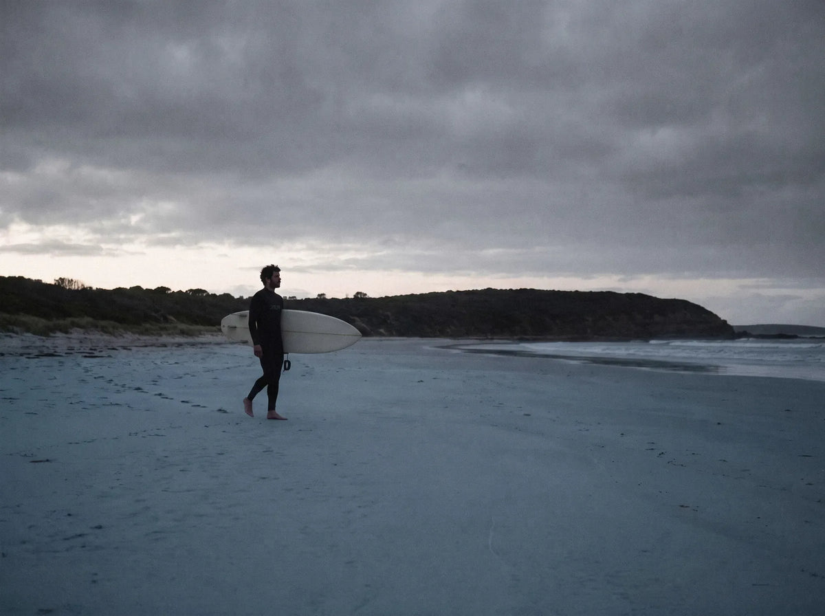 Lone surfer walking an empty beach at dusk with a board under one arm