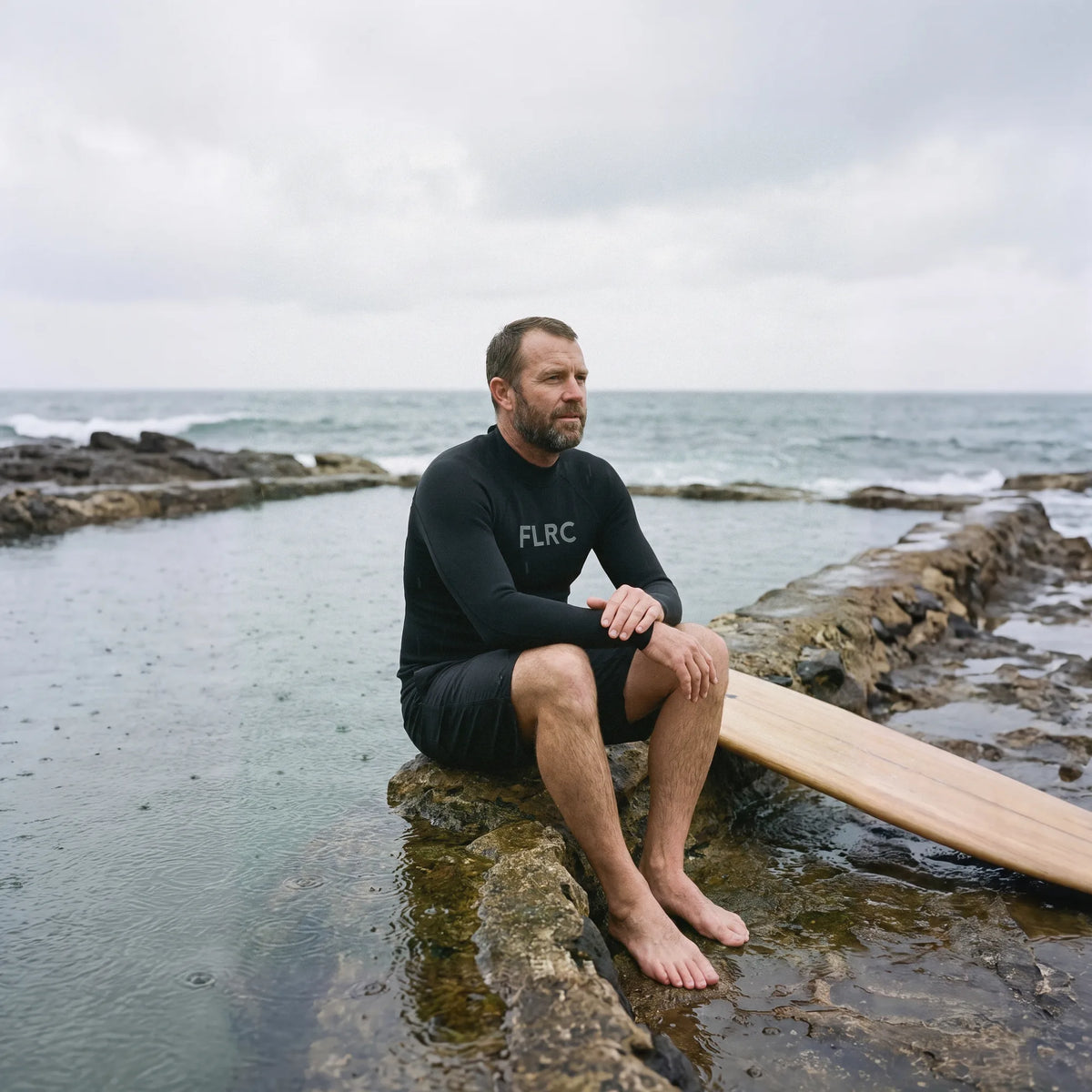 Surfer in the Men's 1.5mm Wetsuit Top V2 sitting on the rocks by an ocean pool, board beside him
